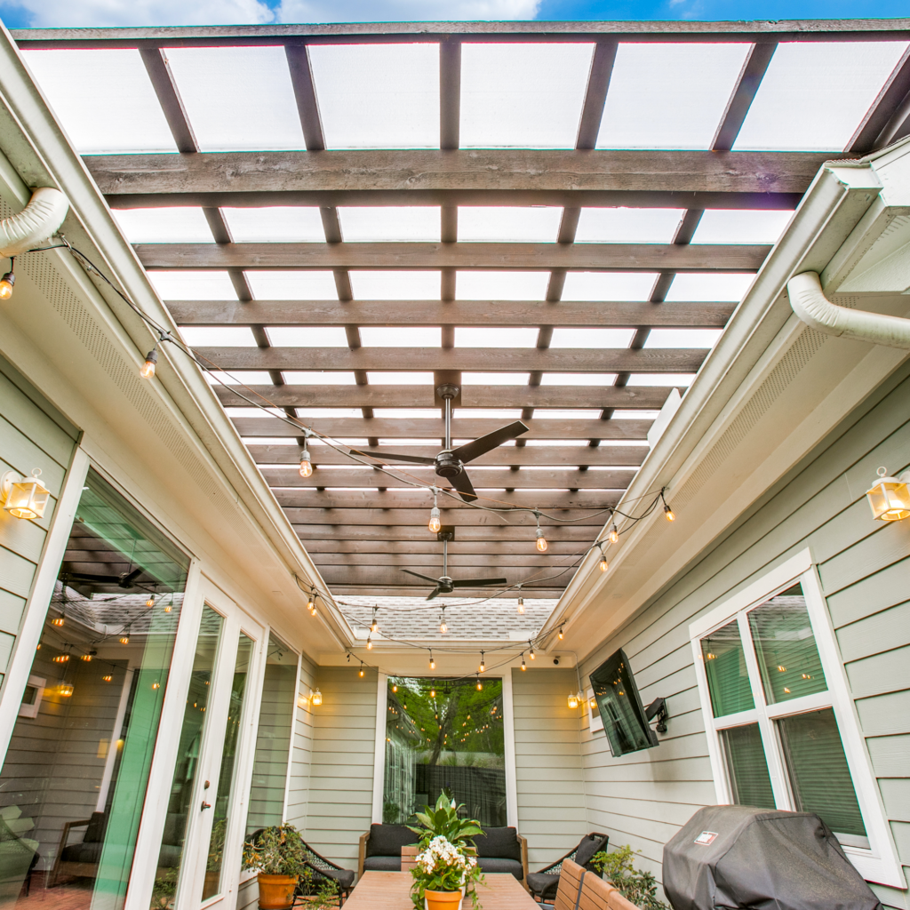 A floating pergola with ceiling fans covered with Hercules White sits over a patio within three sides of a house with windows and a french door. String lights are attached to the gutter system of the house and draped across the patio area.