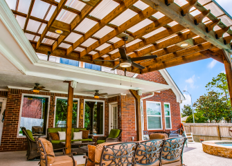 A large pergola with built-in lights and ceiling fan is covered with Skypoly Hercules Bronze and sits over a patio with outdoor furniture near the side of a red brick house with three rectangular windows.
