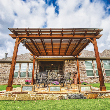 A standalone cedar-stained pergola with hanging lights covered with Hercules Bronze Skypoly sits at the back of a brown and white brick house over a flagstone patio containing a wrought iron outdoor circular table and four chairs and 3 small flower boxes in front.