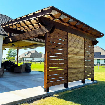 A pergola covered with Skypoly Hercules Bronze with a privacy wall sit on a concrete slab containing outdoor furniture and a grill.