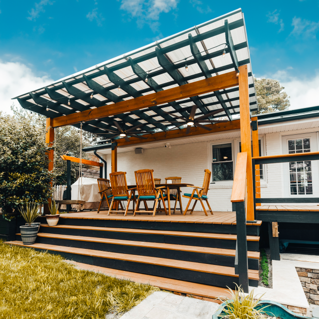 A wooden two-toned pergola covered with Skypoly Hercules White is built on top of a deck. Four steps lead up to the deck where a wooden table and six chairs sit next to the side of the white house near windows and French doors.