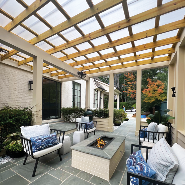 A cream-colored pergola with natural wood joists covered with Skypoly Hercules White is attached to two sides of a white brick house and sits over a patio area which has black metal, white cushioned outdoor furniture, a rectangular fire pit and gray stone tile flooring.