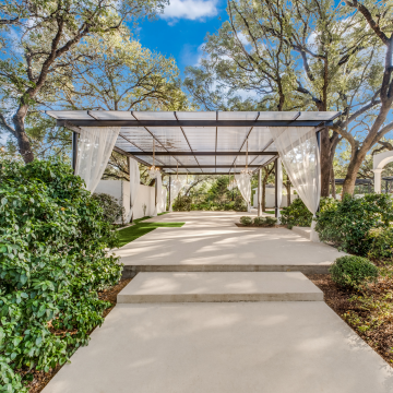 A very large pergola surrounded by majestic Live Oak trees and covered with Hercules White Skypoly sits over a concrete slab with steps at a wedding venue. Crystal chandeliers hang from the ceiling and sheer white curtains are draped at the corners and sides.
