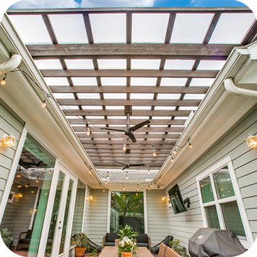 A floating pergola with ceiling fans covered with Hercules White sits over a patio within three sides of a house with windows and a french door. String lights are attached to the gutter system of the house and draped across the patio area.